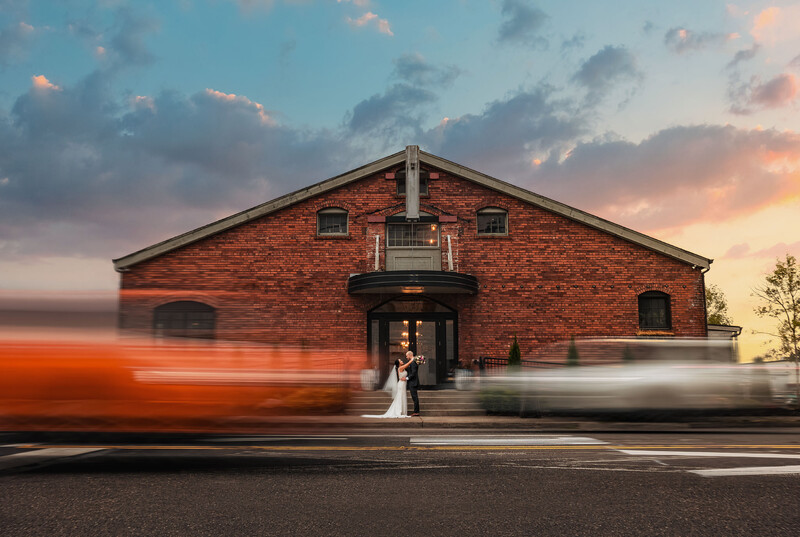 Bride and groom standing in front of a red brick building as blurred vehicles pass by.