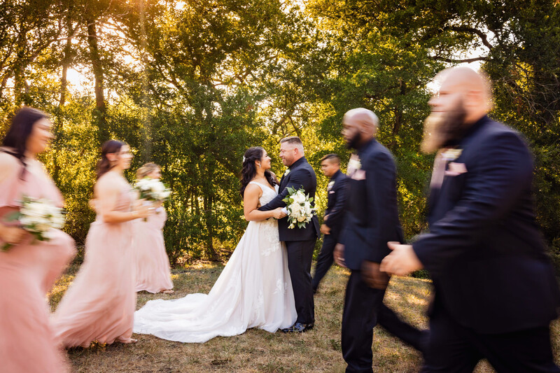 Bride and groom exchanging vows during outdoor wedding ceremony surrounded by wedding party and trees.