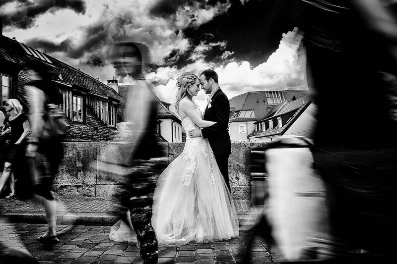 Bride and groom embrace while blurred wedding guests move around them in black and white.