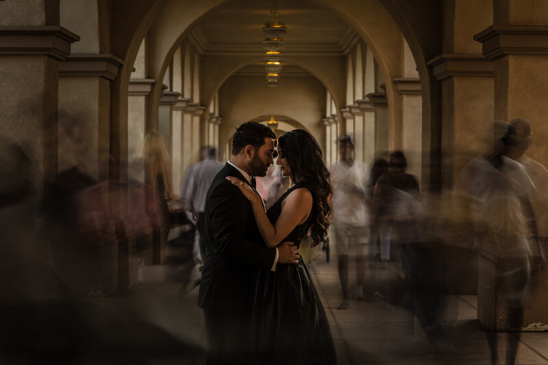 Couple kissing in a classical arched corridor surrounded by blurred figures.