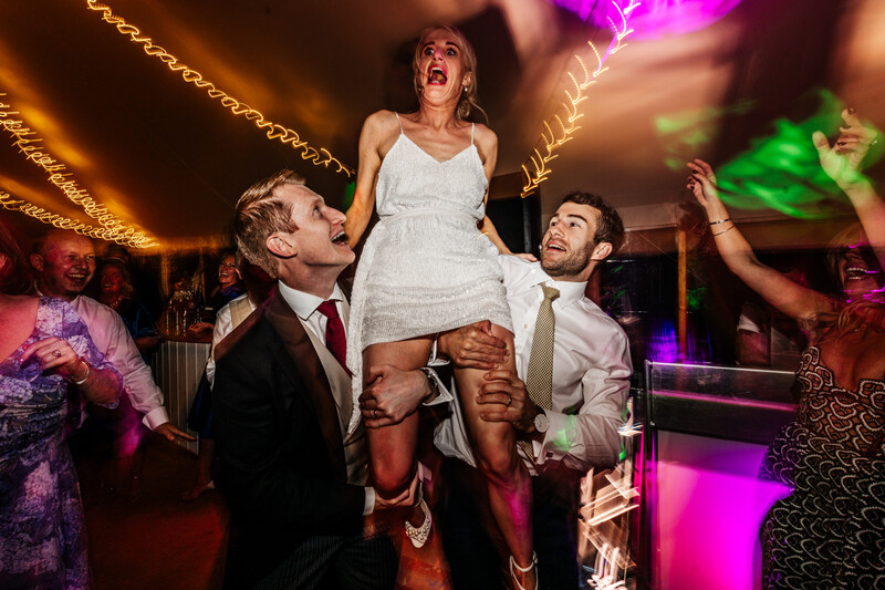 Bride lifted in the air by two men at a wedding reception with colorful ambient lighting.