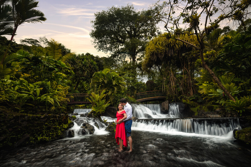 Woman in red dress standing in shallow water before a cascading waterfall surrounded by lush tropical vegetation.