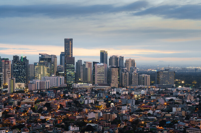 Urban skyline at dusk with illuminated high-rise buildings rising above residential neighborhoods under a cloudy sky.