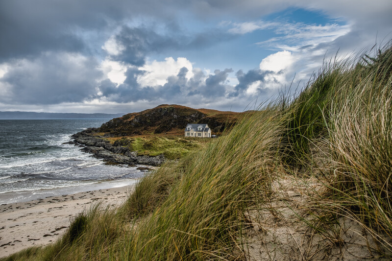 Coastal landscape with marram grass in foreground, sandy beach, white cottage on rocky headland, and dramatic cloudy sky.