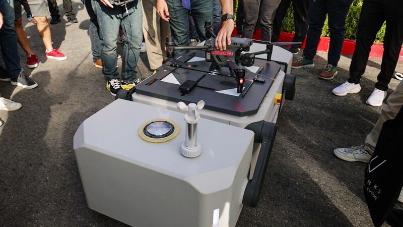 Drone with white and black body positioned on transport case at outdoor demonstration surrounded by spectators.