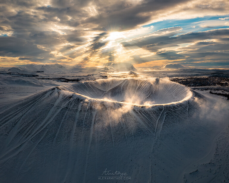 Snow-covered conical volcano with dramatic crepuscular rays breaking through clouds at sunset.