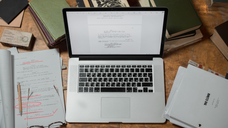 Laptop displaying a document on screen, surrounded by printed pages with red editorial marks and a pen on a wooden desk.