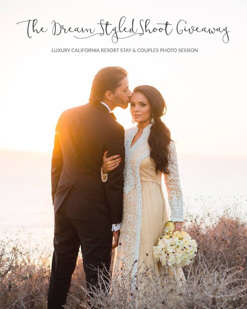 Couple in formal attire embracing outdoors at golden hour with white flowers and dry brush landscape.