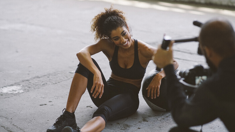 Woman in athletic wear sitting on pavement while someone photographs her with a smartphone.