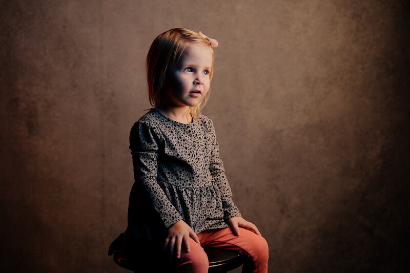 Young child in gray knit sweater and red pants seated against warm brown backdrop.