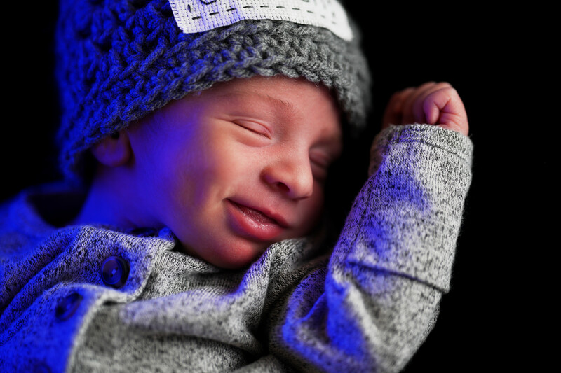 Sleeping newborn baby wearing gray knit hat and blue sweater against black background.
