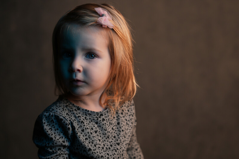Young child with reddish hair wearing a patterned sweater, photographed against a neutral brown background.