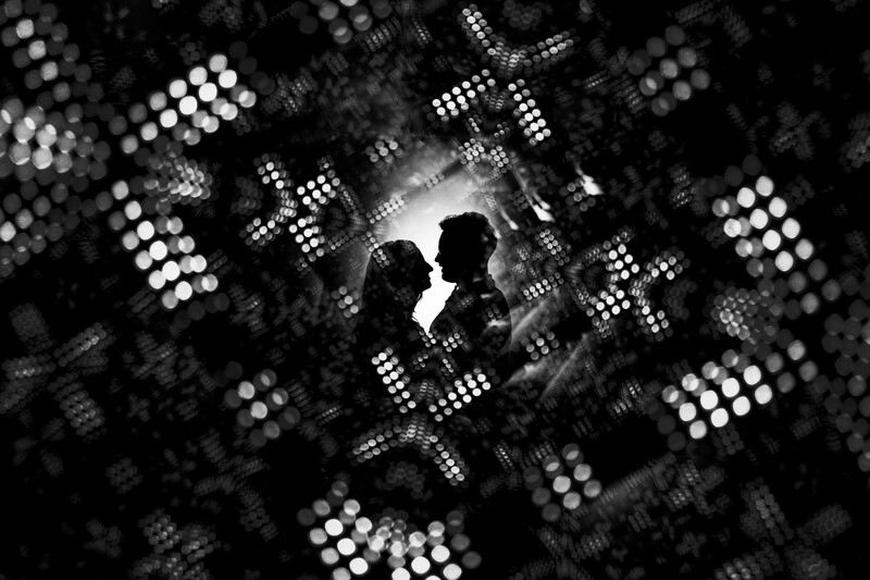 Two silhouetted figures in profile against bright stage lighting and bokeh lights in black and white.