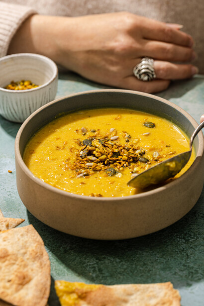 Bowl of golden turmeric soup garnished with seeds, styled with bread and small ingredient bowl.