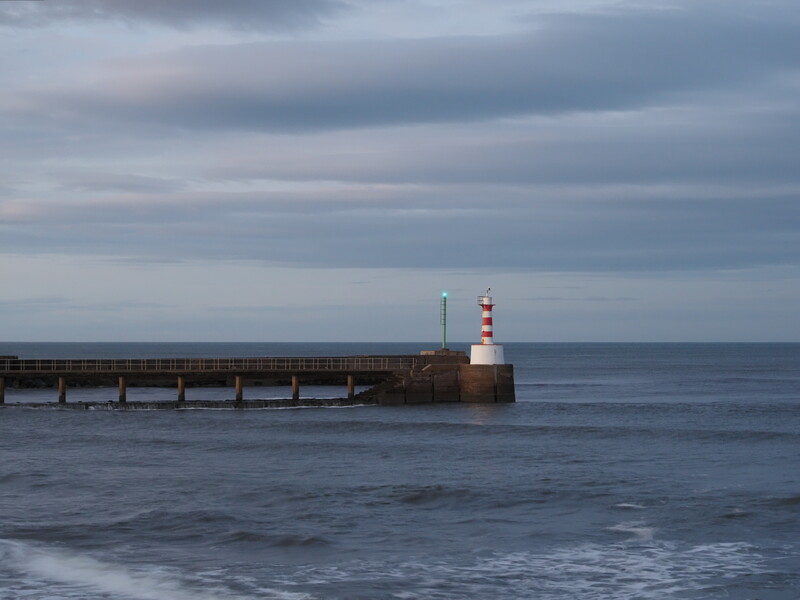 Red and white striped lighthouse on a breakwater extending into calm gray sea under overcast sky.