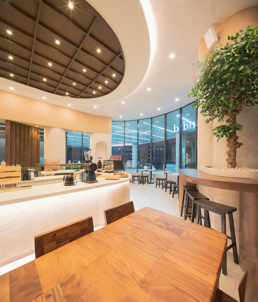 Modern office interior with curved ceiling, wooden dining table in foreground, and floor-to-ceiling windows overlooking city.