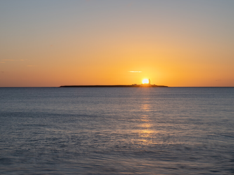 Golden sunset over calm ocean water with a dark landmass silhouetted on the horizon.