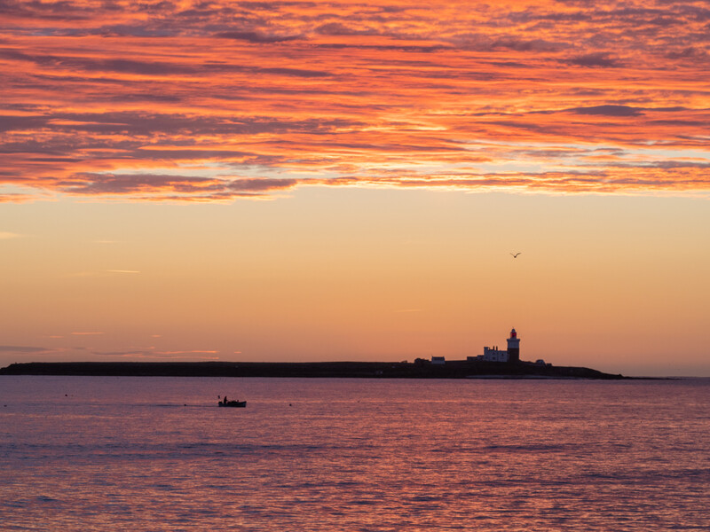 Silhouetted lighthouse on a distant island at sunset with a small boat in the foreground.