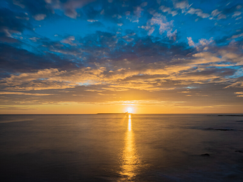 Sun reflected on calm ocean water during golden hour sunset with blue and orange clouds overhead.
