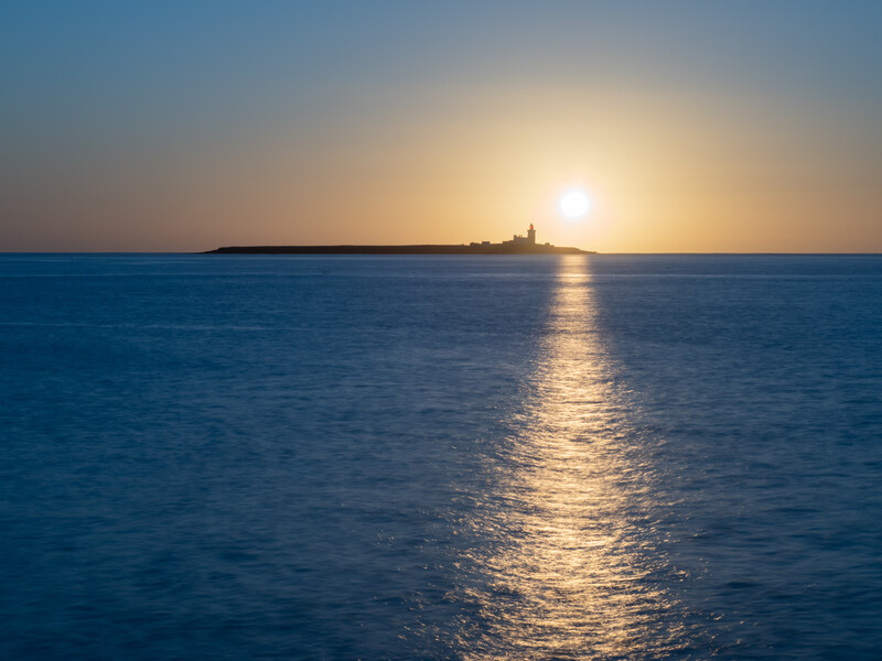 Golden sunlight reflects across calm ocean water at sunset, with a distant silhouette of a coastal structure on the horizon.