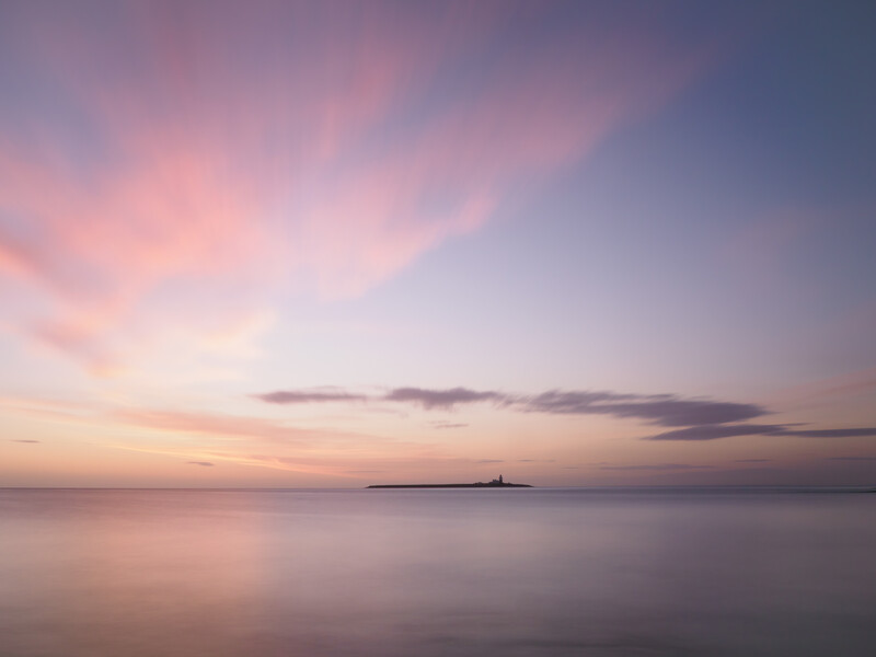 Calm seascape at dusk with a distant silhouetted structure on the horizon beneath soft pink and purple clouds.