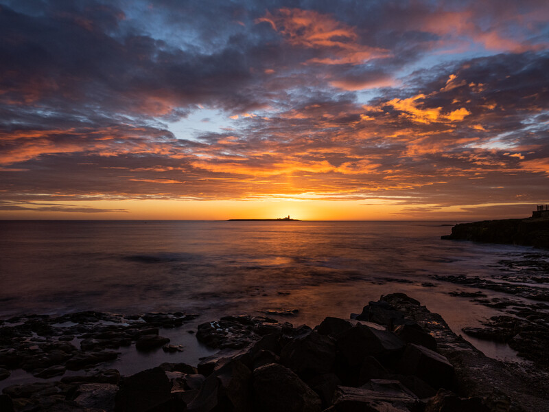 Dramatic sunset over a rocky coastline with golden and orange clouds reflected in still water.