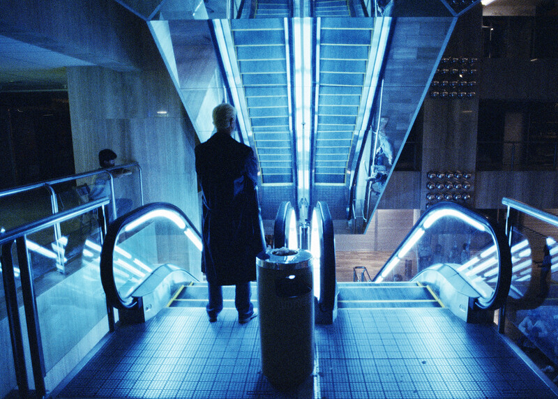 Person standing in a brightly lit subway station with blue-tinted neon lighting and an escalator ahead.