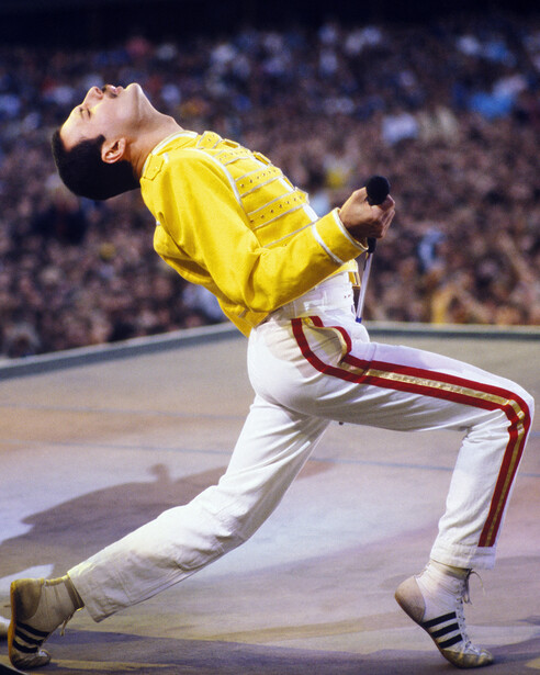 Performer in yellow jacket and white pants arching backward on stage with crowd blurred in background.