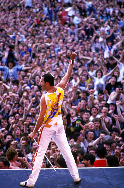 Performer in yellow and white outfit with raised fist standing before massive crowd in arena.