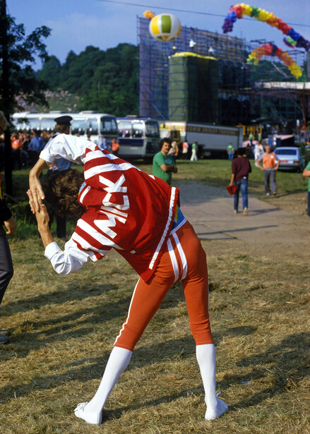 Cheerleader in red and white uniform bending forward on grass with festival stage and crowd in background.