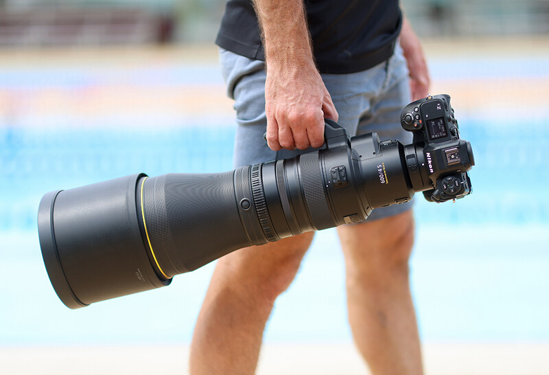 Photographer holding a large telephoto lens with camera body attached near a swimming pool.