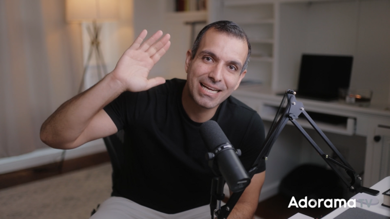 Man waving at camera while speaking into a microphone in a home studio setup.