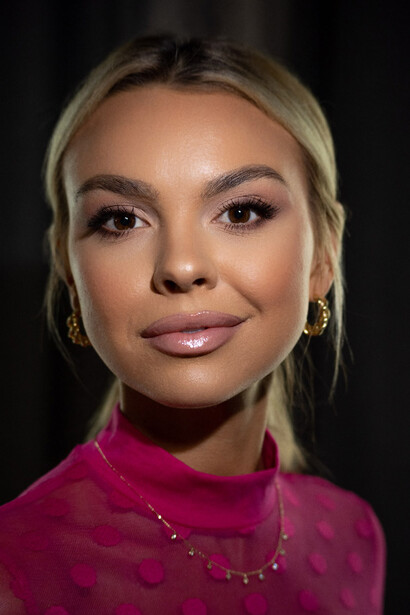 Portrait of a woman with blonde hair wearing a magenta top and delicate chain necklace against a dark background.