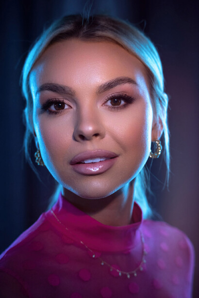 Woman with blonde hair wearing magenta turtleneck against dark background with blue and purple lighting.
