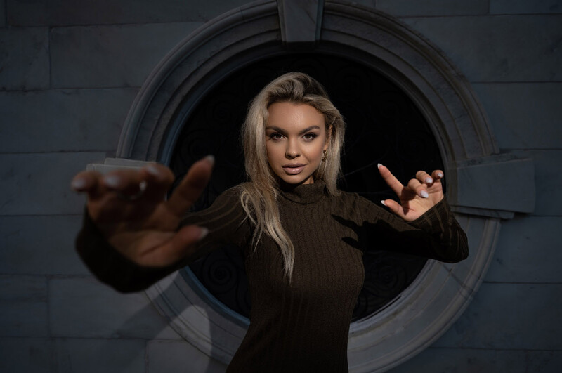 Woman with blonde hair reaching toward camera through circular metallic opening in dark wall.