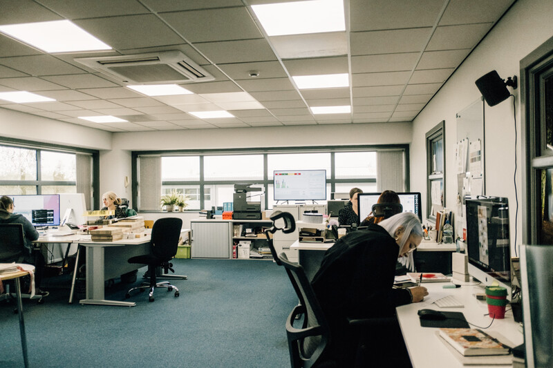 Modern office workspace with multiple employees at desks under fluorescent ceiling lights and large windows.