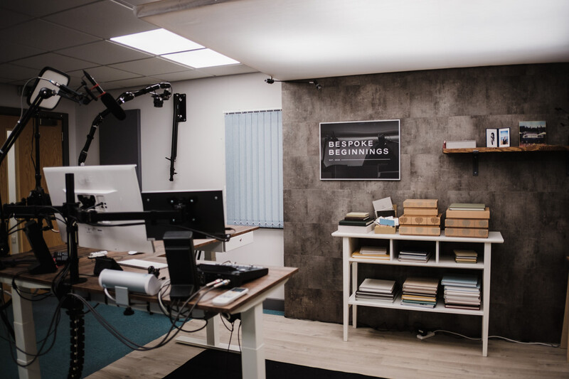 Podcast recording studio with microphone setup, monitor, and wooden shelving against textured gray wall.