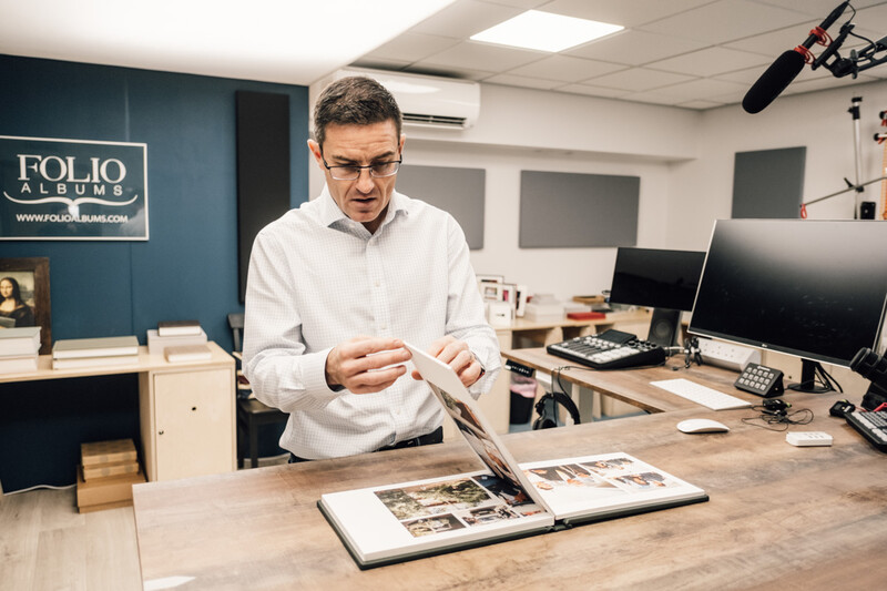 Man in white shirt examining an open portfolio book at a wooden desk in a studio.