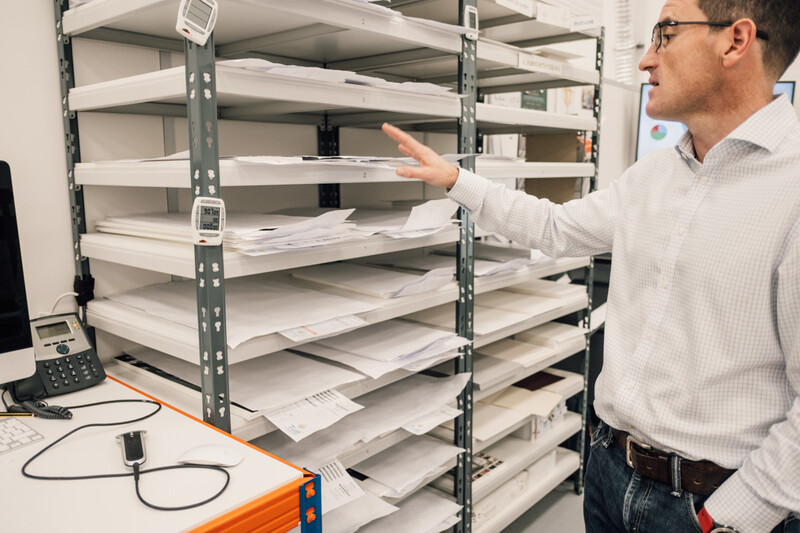 Man in glasses gesturing toward a tall metal shelving unit filled with white papers and documents.