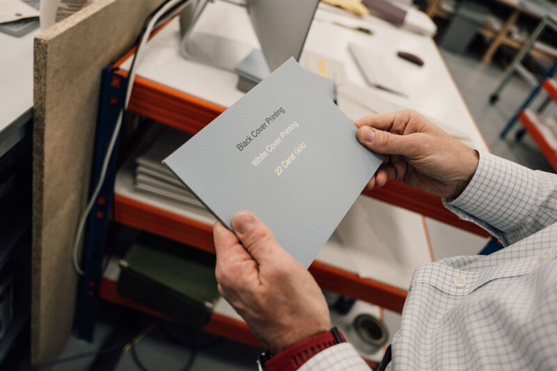Hands holding a gray book or portfolio with gold lettering against a workshop backdrop.