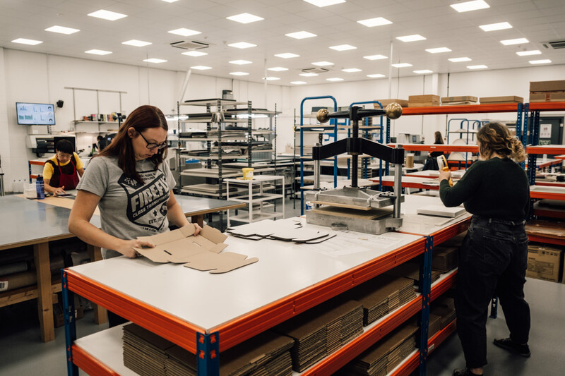 Workers assembling cardboard boxes at industrial work tables in a bright warehouse space.