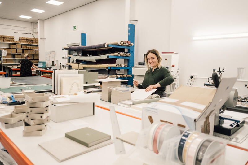 Bookbinding workshop with craftsperson working at table surrounded by materials and equipment.