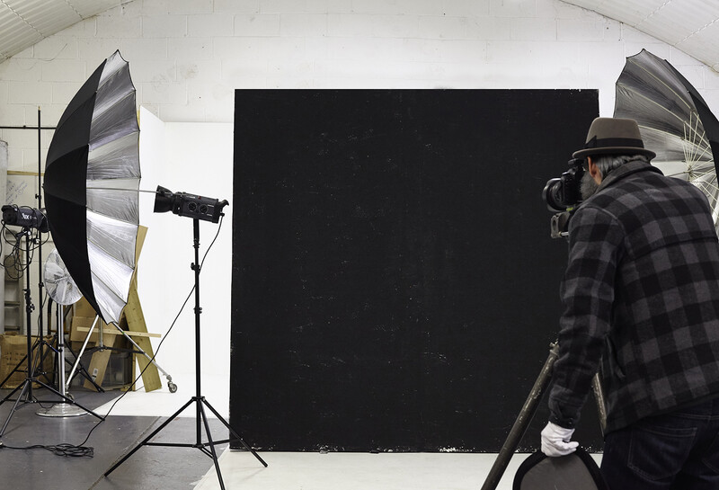 Photographer adjusting camera settings on tripod in studio with large black backdrop and umbrella reflectors.
