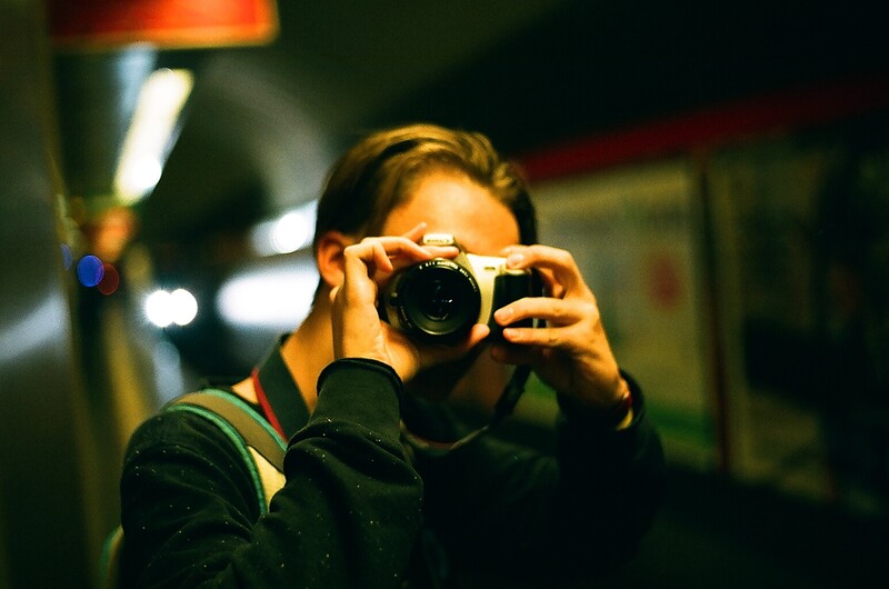 Photographer holding a DSLR camera up to their face while taking a self-portrait in an indoor setting.