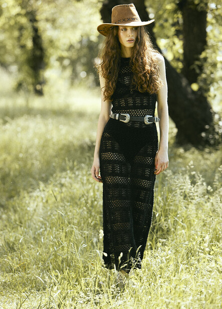 Woman in black lace dress and wide-brimmed hat standing in sunlit grass and trees.