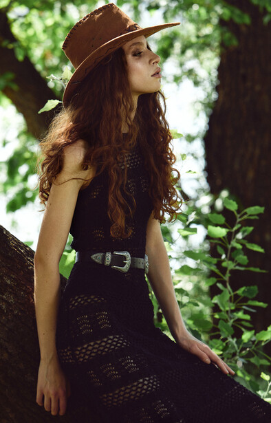 Woman in cowboy hat and black mesh skirt poses among trees with dappled sunlight.