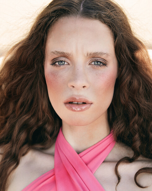 Portrait of woman with long wavy brown hair and pink wrap against neutral background.