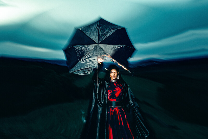 Woman in red dress holding black umbrella under dramatic teal sky and mountains.