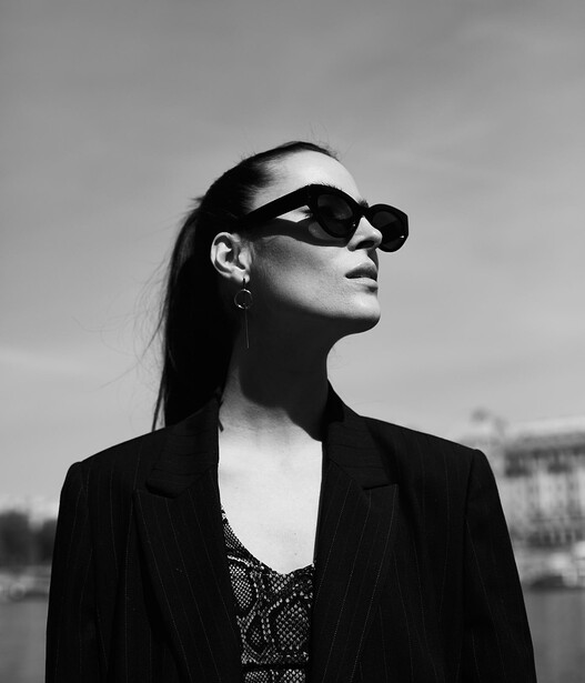 Woman in sunglasses and dark blazer looking upward against overcast sky.