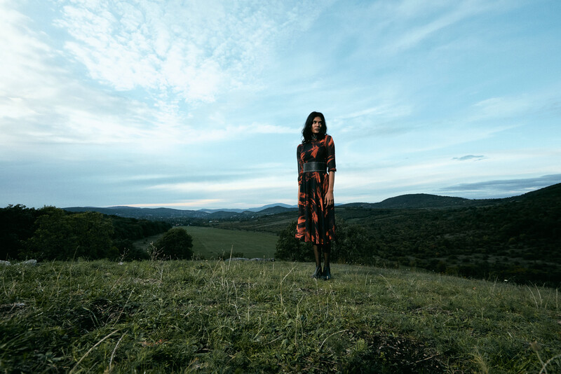Woman in patterned dress standing in open grassland with mountains under blue sky.